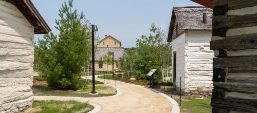A winding path between rustic buildings and trees in a serene outdoor setting.