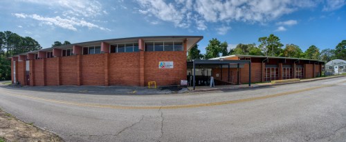 Panoramic view of a brick building with large windows, surrounded by trees and a clear blue sky.