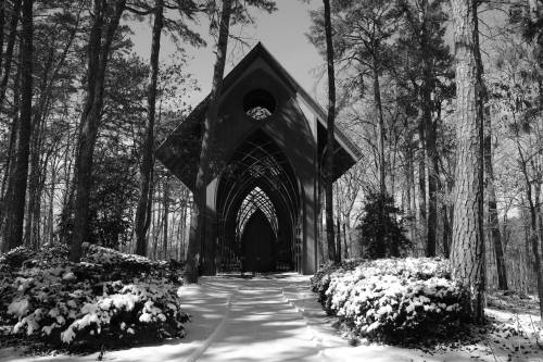 A black and white image of a chapel surrounded by snow-covered trees and a path leading to its entrance.