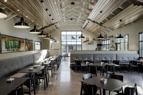 Modern restaurant interior with high ceilings, gray booths, black tables, and large windows letting in natural light.