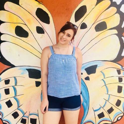 A woman poses in front of a colorful butterfly mural, wearing a blue top and shorts, smiling at the camera.