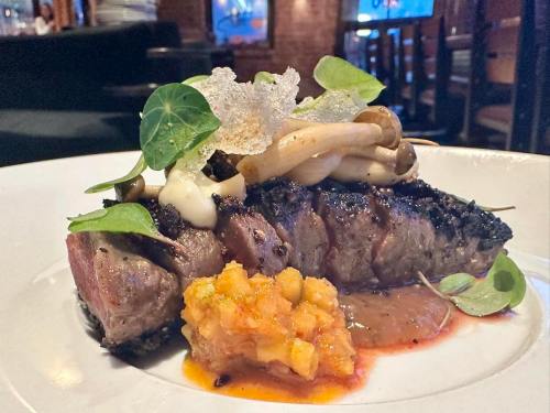 A beautifully plated steak topped with mushrooms, greens, and a colorful sauce on a white dish.