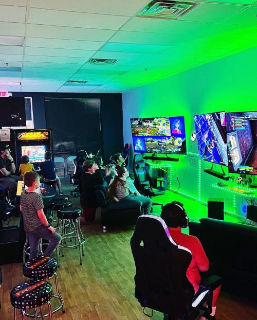 A gaming lounge with people playing video games on large screens, surrounded by colorful lights and arcade machines.