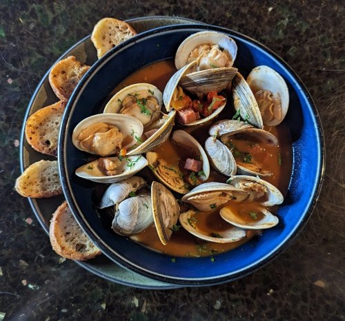 A blue bowl filled with clams in broth, garnished with herbs, accompanied by slices of toasted bread.