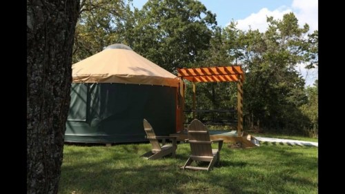 A yurt surrounded by trees, featuring a wooden deck and two Adirondack chairs on a grassy area.