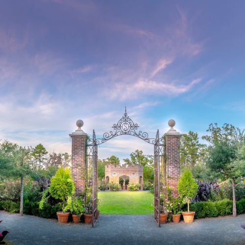 A picturesque garden entrance with wrought iron gates, leading to a lush green lawn surrounded by trees and plants.