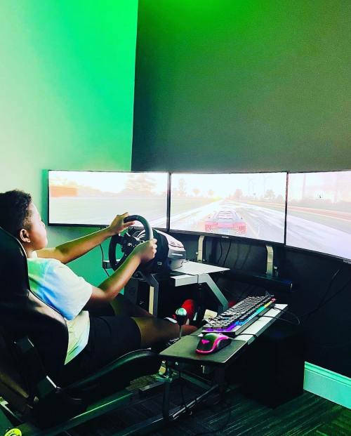 A child sits in a gaming chair, focused on racing on three monitors with a steering wheel setup.