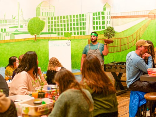 A person in overalls holds a plant while speaking to an audience in a colorful, mural-decorated room.