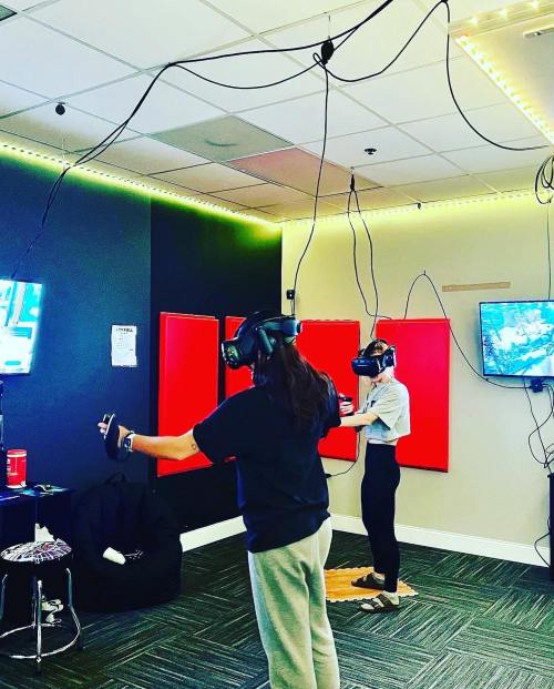 Two people wearing VR headsets in a room with screens and cables, engaged in a virtual reality experience.