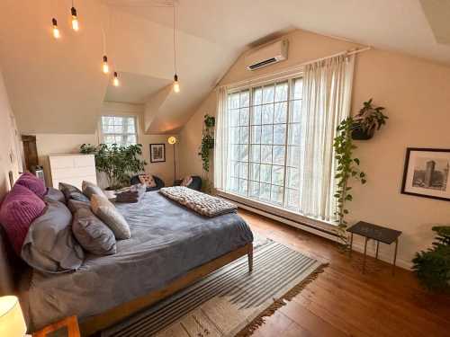 Cozy bedroom with a large window, plants, a bed with purple pillows, and warm lighting.