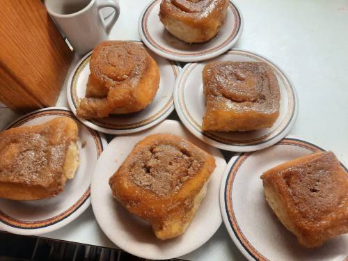 A collection of cinnamon rolls on plates, some with icing, arranged on a table with a small cup in the background.