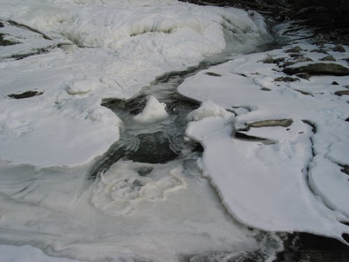 A frozen river with thick layers of ice and snow, creating a textured, white landscape.