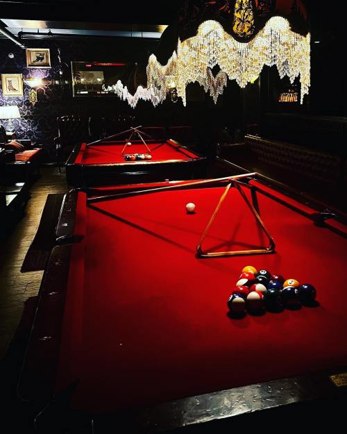 Two pool tables with red felt, one with a triangle of balls, set in a dimly lit, stylish bar environment.