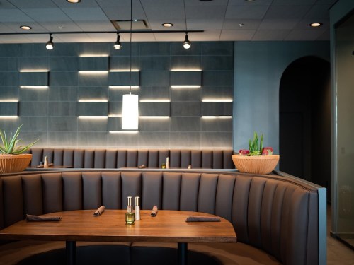 Cozy restaurant interior with a curved booth, wooden table, and modern lighting, featuring plants in decorative bowls.