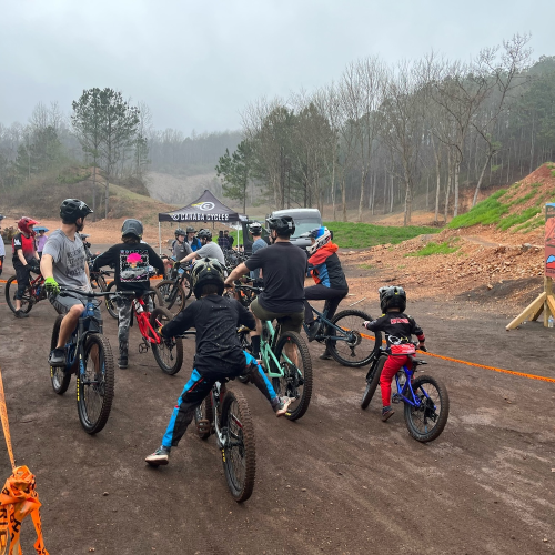 A group of cyclists prepares to ride on a dirt trail in a wooded area, with a cloudy sky overhead.