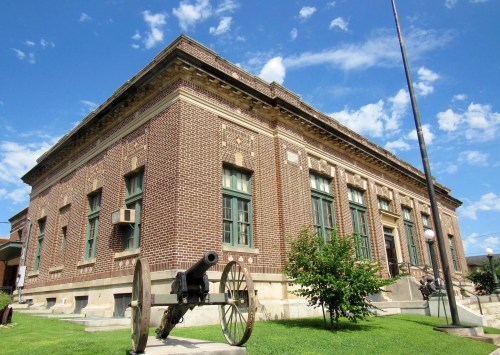 Historic brick building with large windows, surrounded by green grass and a cannon in the foreground under a blue sky.