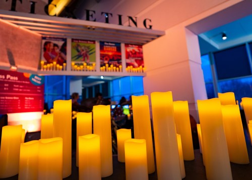 A close-up of glowing candles on a table, with colorful movie posters in the background and a soft blue light.