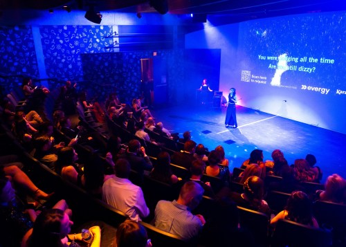 A performer on stage addresses an audience in a dimly lit theater, with colorful lighting and a large screen behind.