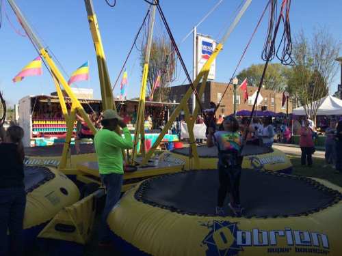 A child jumps on a bungee trampoline at a fair, with colorful flags and booths in the background.