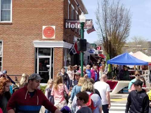 A busy street scene with people walking, shops, and tents set up for a market day under clear blue skies.