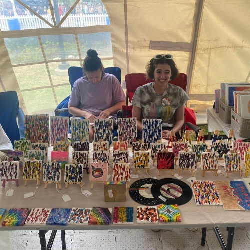 A colorful display of artwork on tables inside a tent, with two people sitting behind the art.