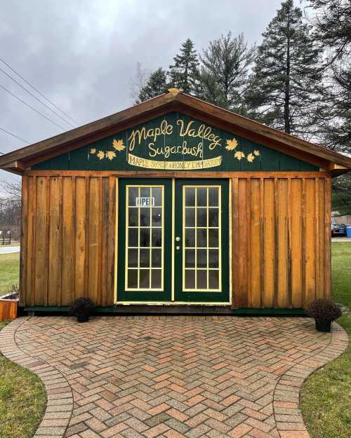 A wooden sugar shack with green accents, featuring double doors and a sign that reads "Maple Valley Sugarbush."