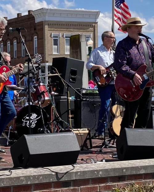 A band performs outdoors with guitars and drums, set against a backdrop of buildings and an American flag.