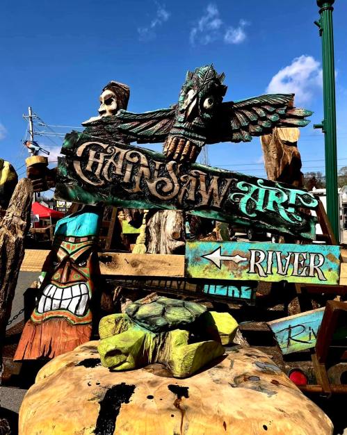 Colorful wooden sculptures and signs, including an owl and a tiki figure, displayed at an outdoor event.
