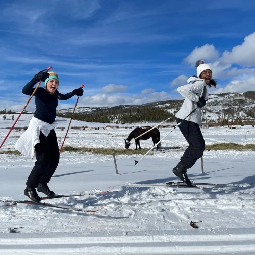 Two women joyfully cross-country skiing in a snowy landscape with a horse in the background under a blue sky.