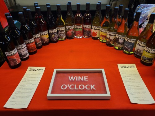 A table with various wine bottles arranged in a circle, featuring a sign that reads "WINE O'CLOCK" and menus.