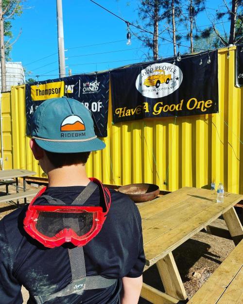A child wearing a cap and goggles stands in front of a colorful outdoor space with banners and picnic tables.