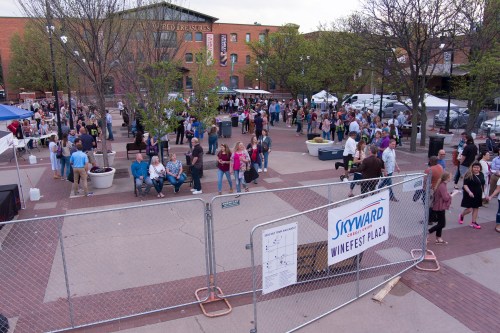 A lively outdoor event with crowds, tents, and trees in a plaza, featuring people socializing and enjoying the atmosphere.