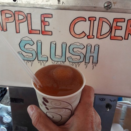 A hand holds a cup of apple cider slush in front of a colorful sign displaying the drink's name.
