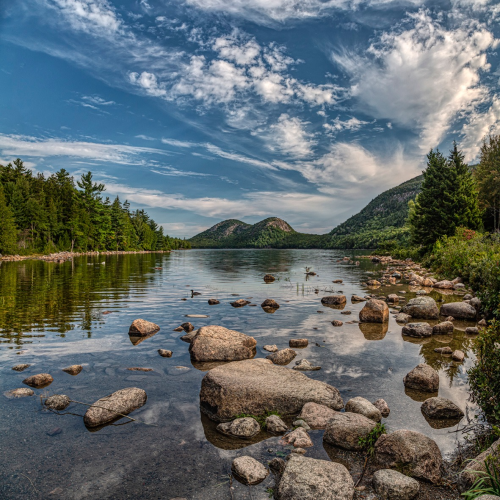 A serene lake surrounded by lush greenery and rocky shores, under a blue sky with scattered clouds.