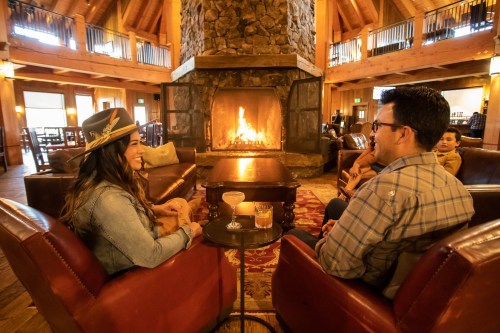 A cozy lodge interior with a couple enjoying drinks by a stone fireplace, surrounded by warm wooden decor.