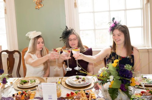 Three women in elegant attire toast with cocktails at a beautifully set table adorned with flowers and gourmet food.