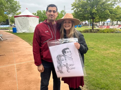 A man and woman pose outdoors, holding a caricature drawing of themselves, with trees and tents in the background.