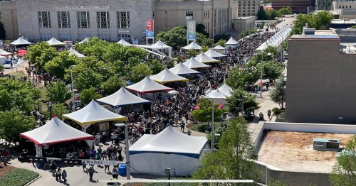 Aerial view of a large crowd at an outdoor event with numerous tents and trees lining the area.