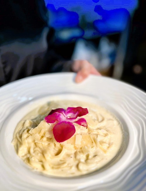 A plate of creamy pasta topped with cheese and edible flowers, held by a person in a dimly lit setting.
