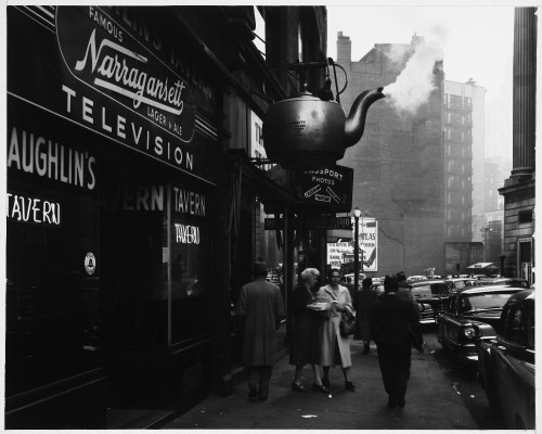 A vintage street scene featuring a large teapot sign, pedestrians, and classic cars near a tavern.