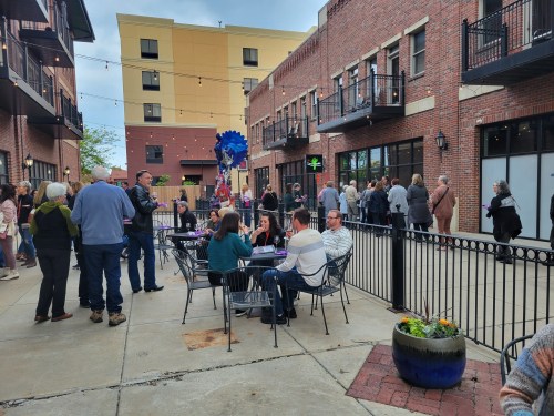 A lively outdoor gathering with people socializing at tables near a brick building, surrounded by greenery and string lights.