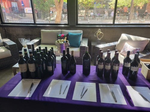 A table with various wine bottles and pens, set up in a bright space with seating and greenery visible outside.
