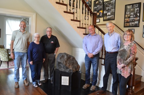 A group of six people stands beside a large rock display in a home, with a staircase and photo wall in the background.