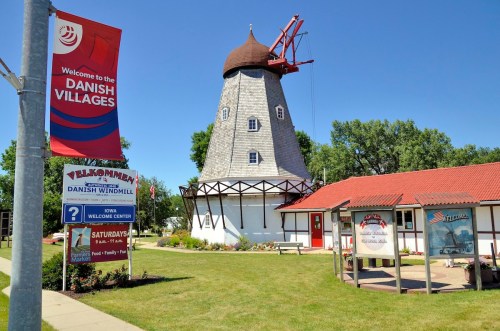 A traditional windmill in a village setting, with a welcome sign and vibrant landscaping under a clear blue sky.