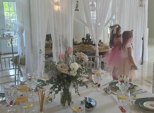 A beautifully set table with flowers and drinks, with two children in fairy costumes playing in the background.