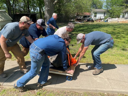 A group of six men work together to move a large rock on a wooden platform with wheels in a grassy area.