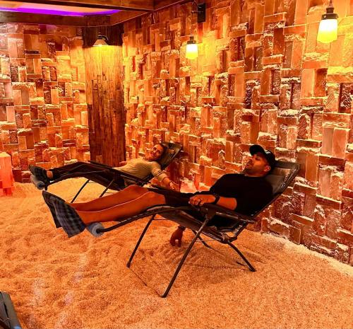 Two people relax on lounge chairs in a cozy room with a salt brick wall and soft lighting.