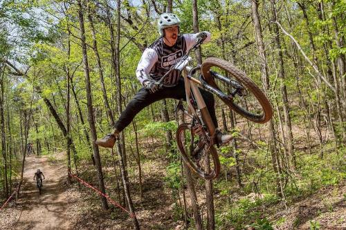 A mountain biker performs a jump over a trail in a wooded area, showcasing excitement and skill.