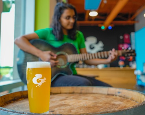 A glass of beer sits on a barrel in the foreground, while a person plays guitar in the background.