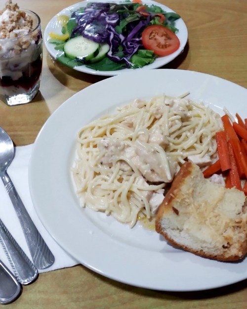 A plate of creamy chicken pasta, carrots, and bread, with a side salad and a dessert glass on a table.